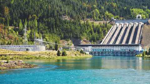 A hydroelectric dam, with blue-green water in the foreground and pine forests on hills behind it.