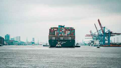 A container ship moving through a busy port, with a city in the background.