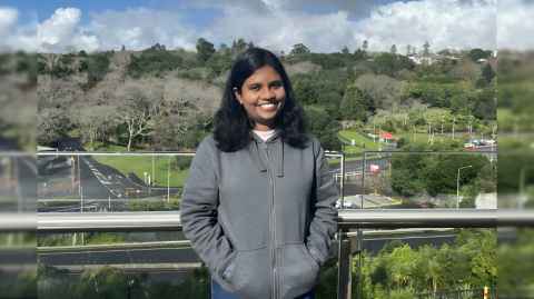 A smiling young woman in a hoodie, standing on a balcony with the Auckland Domain in the background.