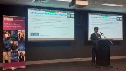 A man standing at a lectern with slides headed ' Challenges and difficulties to negotiate' displayed on screens behind him.