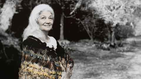 A grey-haired woman wearing a korowai, with trees in the background.