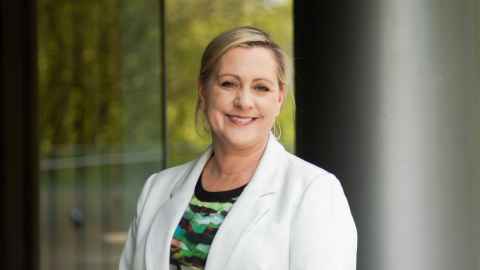 A blonde woman in a white blazer and multicoloured shirt standing in front of a glass window.