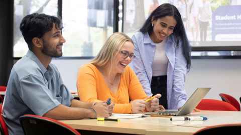 Three students sitting at a table all looking at one laptop and smiling.