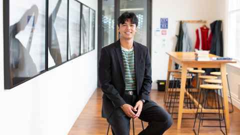 A man with short dark hair sitting on a high stool in a modern office, wearing a dark suit and colourful t-shirt, smiling.
