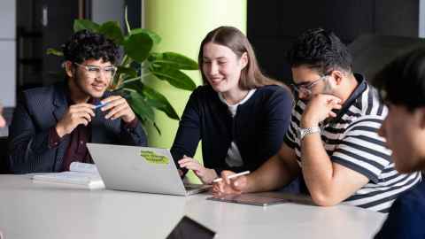 Group of four students sitting at a table looking at a laptop.
