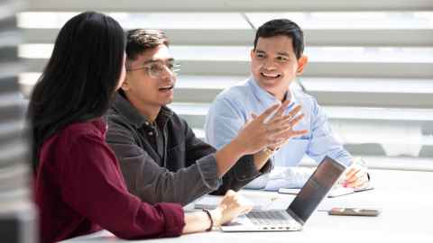 Three students talking at a table and looking at a laptop.