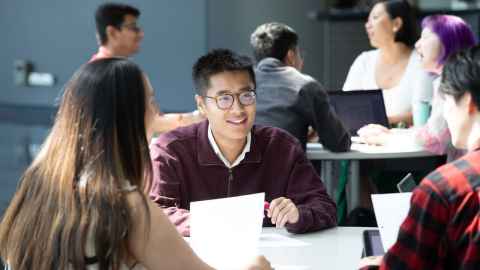 Man with short dark hair and glasses, sitting at a table with other people talking. 