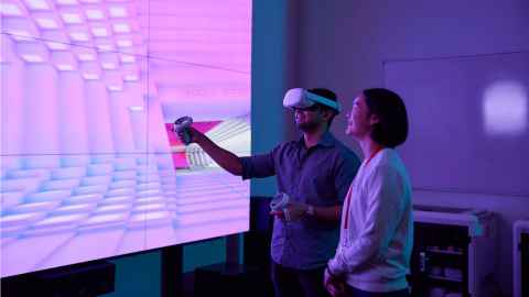 Two students in front of a large screen, with one wearing a VR head set