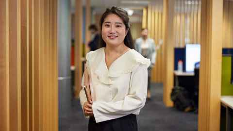 Postgraduate student standing in the hallway, holding a notebook and smiling.