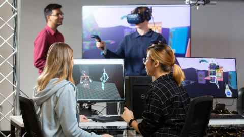 2 people on computer, 2 people in background using virtual reality headsets and equipment