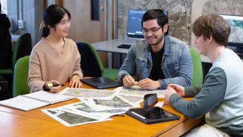 Three students sitting and talking at a table, with maps and paper in front of them.
