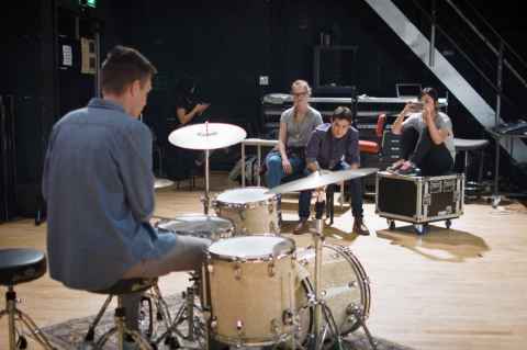 Close up person playing drums, as other students watch them, sitting near musical equipment