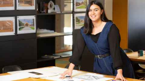 Architecture graduate wearing a denim dress and black shirt smiles while standing and leaning on desk in the architectural firm where she works, models and sketches are on the walls behind her. 