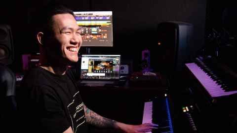 Music graduate sitting in front of keyboards and computer in his music production studio, smiling into the distance.