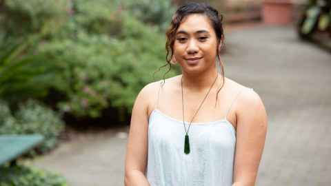 Dance student Chas standing outside in courtyard with plants, wearing white dress and pounamu