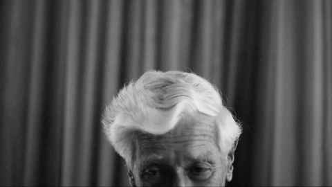 Elderly person's face in front of curtain, in black and white.