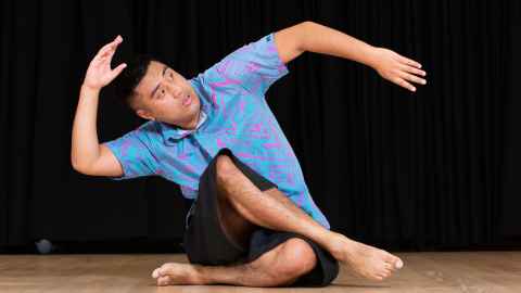 Xavier Breed wearing a colourful cultural clothing, sitting down, legs crossed, arm moving in the air, mid dance pose.