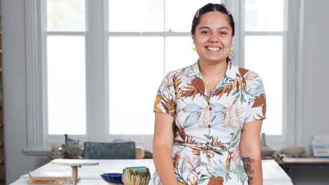 Fine Arts graduate and artist Ashleigh Taupaki smiling and sitting on a table in her studio in Auckland