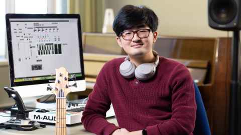 Music student Kihoon sitting in front of computer, wearing headphones around his neck and a guitar sitting beside him.
