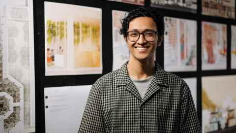 Photo by David St George. Rishav Sarmah stands in front of his NZIA commended student work which is pinned to boards behind him, he smiles to camera.