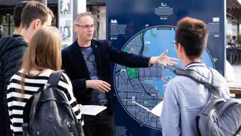 Associate Professor Lee Beattie standing in front of public sign in Wynyard Quarter, Auckland. His arms are outstretched and he is talking with three students, whose backs face the camera.