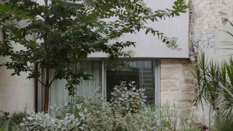 A steel/stone house nestled behind bushes and trees