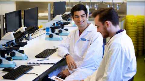 Two young men in lab coats, sitting in front of microscopes and computers.
