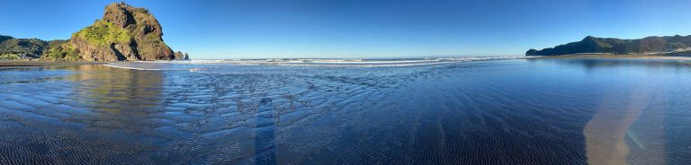 Piha: panorama of Lion Rock to North Piha headland with light blue sky and waves in between