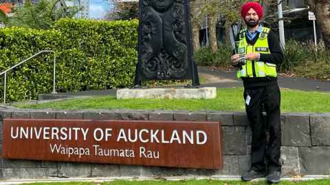 Ajmer in his high-vis vest and red turban standing by a University of Auckland sign and carving