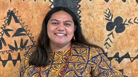 Dantzel Rose Tiakia smiling, wearing a tapa print shirt against a tapa cloth