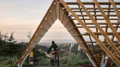 Foreground: timber framing of a building, looking throgh an A-frame, which frames a cyclist on a ridge. Background is land and sky