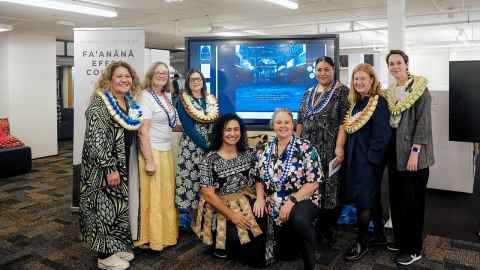 Photo of a group of women standing in front of a large tv screen. The women are all wearing Pacific cultural clothing and jewellery. 