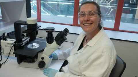 Photo of Dr Molly Swanson in her lab coat and lab PPE. In front of her is an electric microscope. 