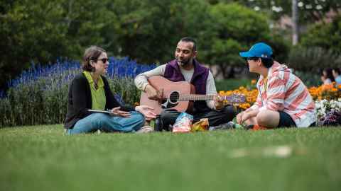 Photo of people sitting in a park. One person is holding a guitar while the other three are looking at him. 