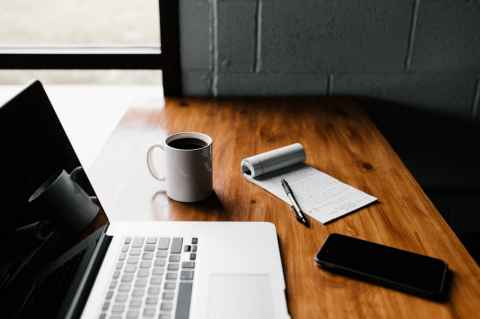 laptop on a desk with coffee, a phone and a pen and paper