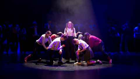 A group of people bowing to a young woman in pigtails on a mostly dark stage, with a spotlight in the centre.