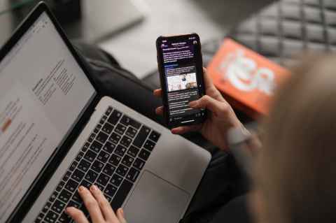 Person typing on a laptop while browsing on a mobile phone.