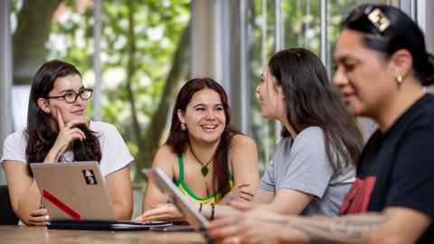 Students sitting around a table, talking and looking at laptops.
