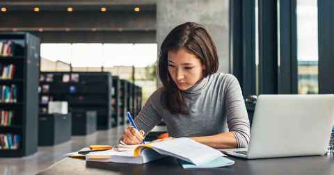 Woman sitting as desk in a library, writing in a book with a laptop open next to her.