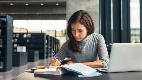 Female student taking notes from a book at library. Young Asian woman sitting at table doing assignments in college library.