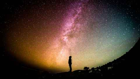 Silhouette of a person standing on a rocky hill under a vivid star-filled night sky with the Milky Way stretching overhead in pink, purple, and green hues.