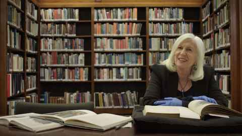 Dame Anne Salmond sitting at a desk with open books before her and bookcases in the background.
