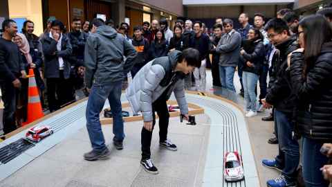 Two students in the centre of a model car race track racing their remote control cars. Spectators look on from outside the track.