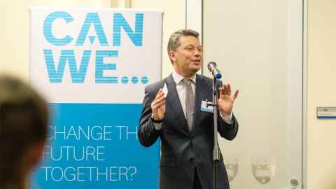 Person standing at a microphone in front of a banner that reads "Can we... change the future together?"