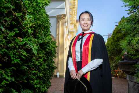Jenny Wang, wearing academic regalia, standing in the gardens outside Old Government House