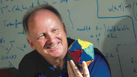 Distinguished Professor Marston Conder holding a model of a geometric shape in front of a whiteboard with mathematical formulae.