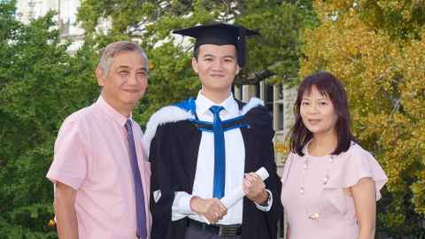 Tristan Pang, wearing academic regalia, standing between his with parents Thomas and Elaine Pang in the grounds behind the ClockTower.