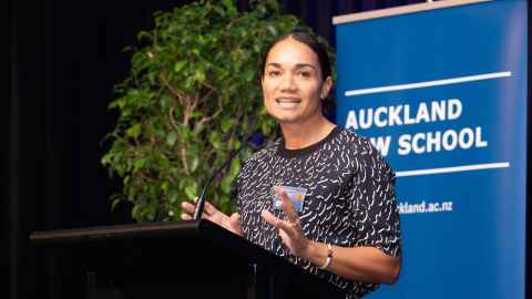 Tiana Epati speaking at a podium in front of a banner that reads "Auckland Law School"