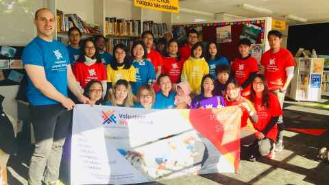 A group of volunteers in a school library. They are wearing coloured T-shirts with Volunteer Impact Week logos, and holding a Volunteer Impact Week banner.