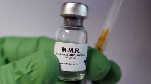 A small clear glass vial labelled "M.M.R. measles, mumps, rubella vaccine" being held in a hand wearing bright green medical gloves together with a syringe.